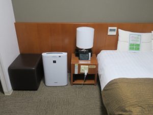 ホテルドーミーイン稚内 ダブルルーム 枕元 スツールと加湿空気清浄機、サイドテーブル、目覚まし時計、エアコンと室内のスイッチがあります