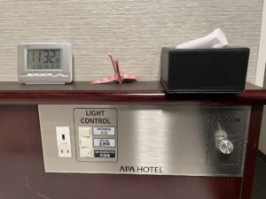 アパホテル 千葉中央駅前 シングルルーム 枕元 目覚まし時計とティッシュ、照明とエアコンのスイッチ、ACコンセント、USBポートがあります