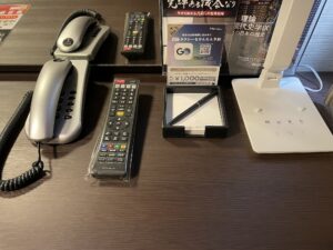 アパホテル 千葉中央駅前 シングルルーム デスク上 電話機とテレビのリモコン、メモ帳、明かり