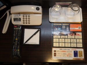 アパホテル 仙台駅五橋 シングルルーム デスク上 電話機とテレビのリモコン、LANポート、コップ