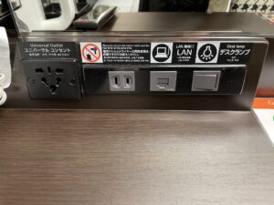 アパホテル 東新宿歌舞伎町タワー スタンダードルーム デスク上 ACコンセントとLANポート、デスクランプのスイッチがあります