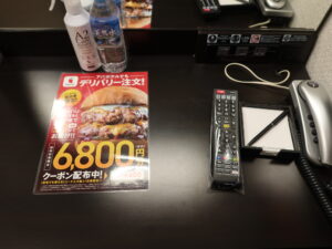 アパホテル 名古屋丸の内駅前 シングルルーム デスク上 テレビのリモコン、電話機、明かりのスイッチ、LANポート、ACコンセントがあります